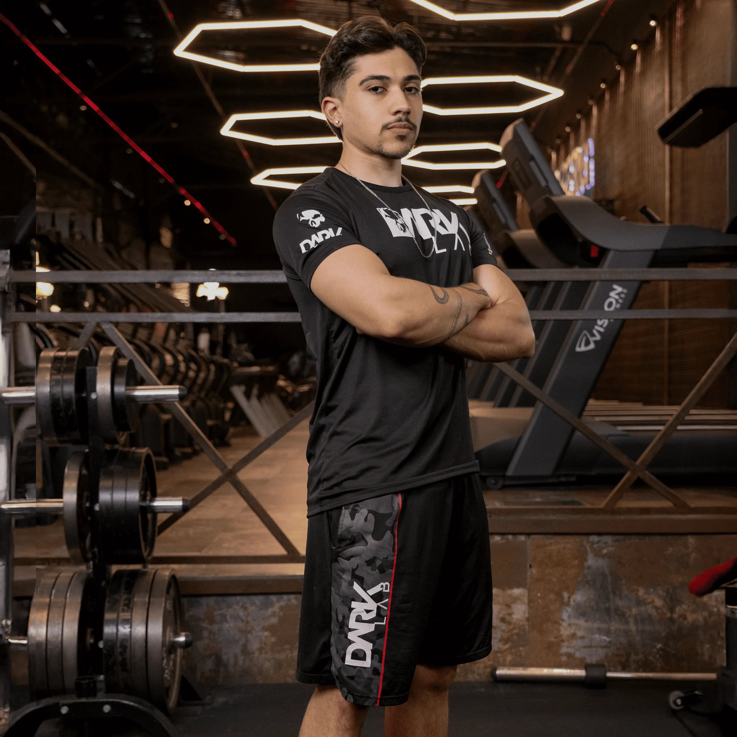 Homem na academia com braços cruzados de perfil usando a bermuda masculina Dark Lab que tem o logo da marca na parte lateral