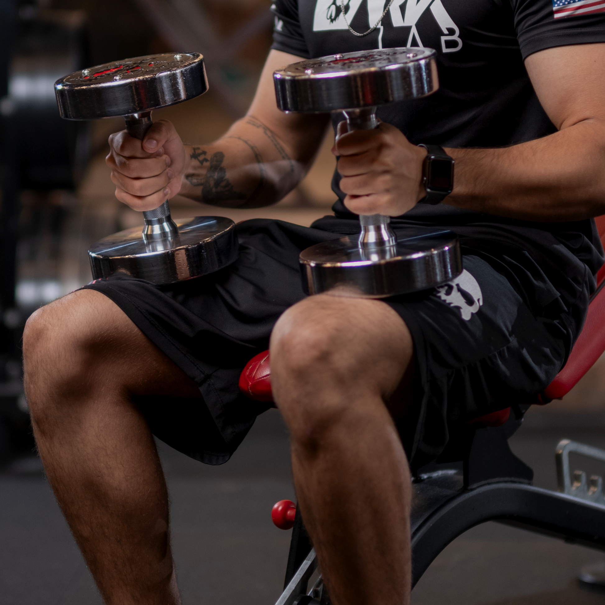 homem sentado em aparelho na academia com foco em suas mãos segurando pesos de academia sobre as pernas e sobre a bermuda masculina da marca Dark Lab