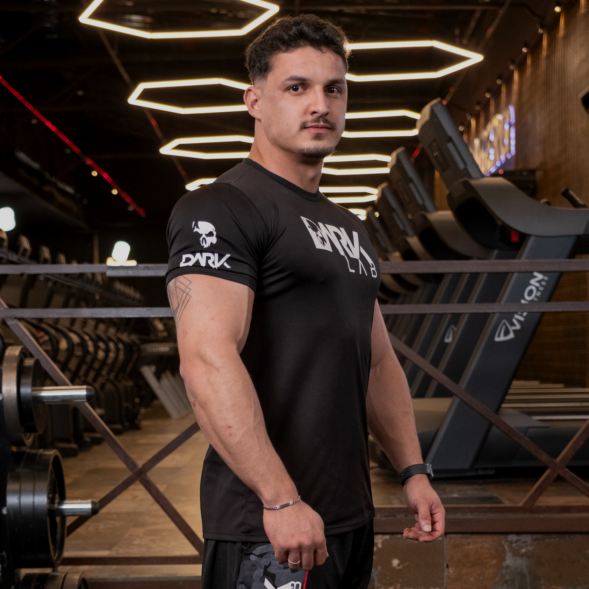 Homem de perfil na academia usando Camiseta de Caveira para Treino da Dark Lab que tem a logo da marca na região frontal e a caveira ícone da marca na manga