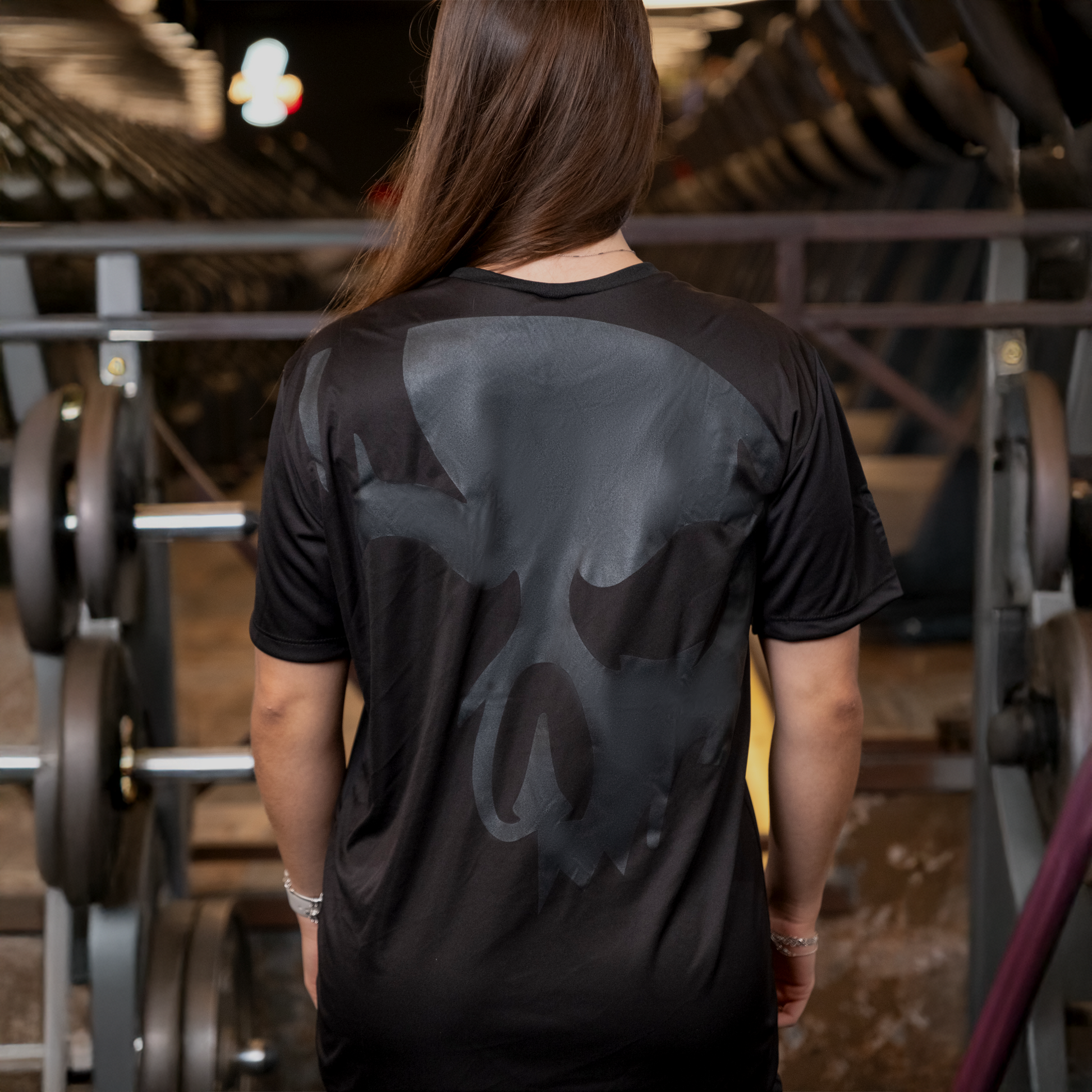 Mulher de costas na academia usando camiseta Dry-fit Preta de caveira da Dark Lab mostrando a parte de trás da camiseta que contém a caveira ícone da marca 