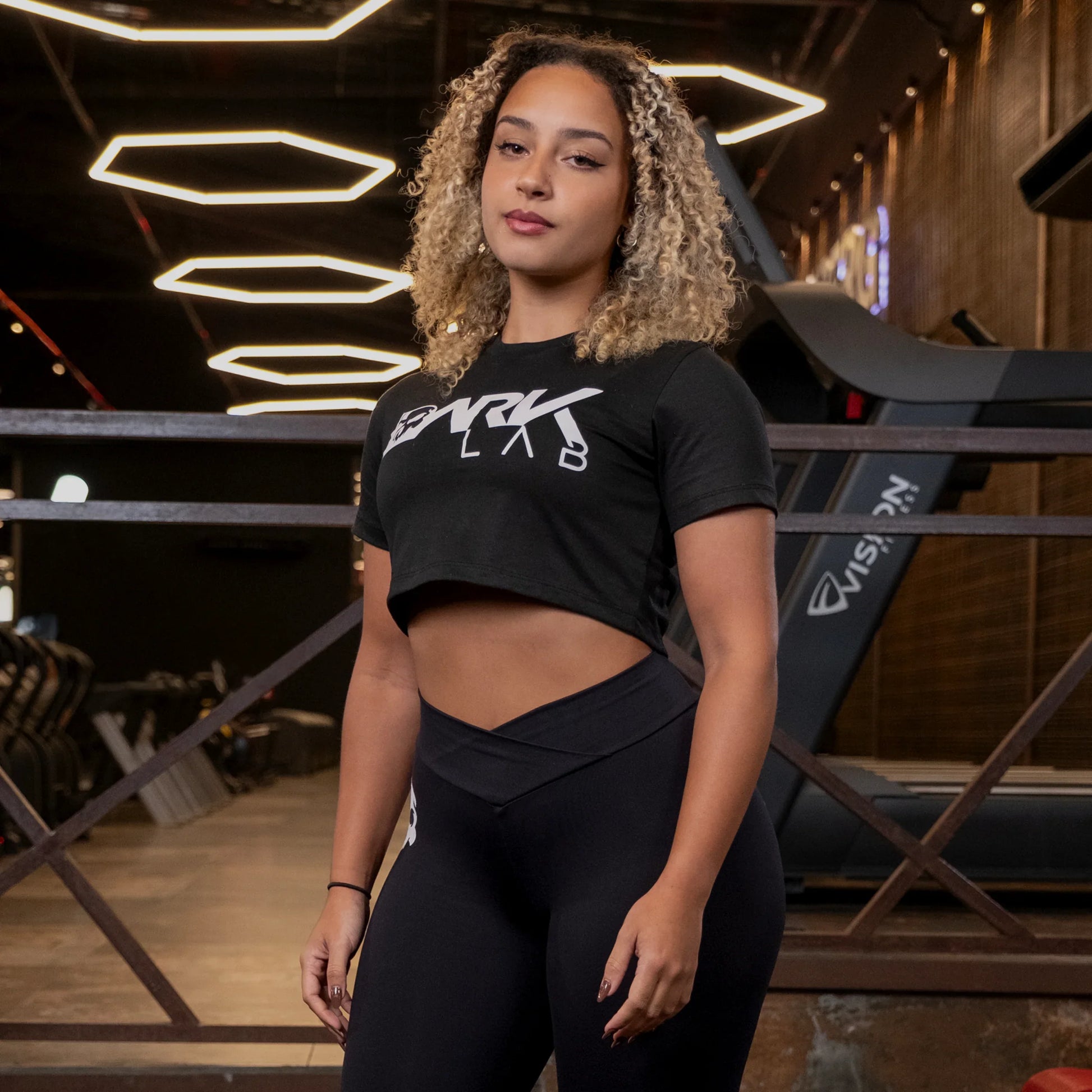 Mulher com fundo de academia usando o Cropped Fitnes preto da Dark Lab