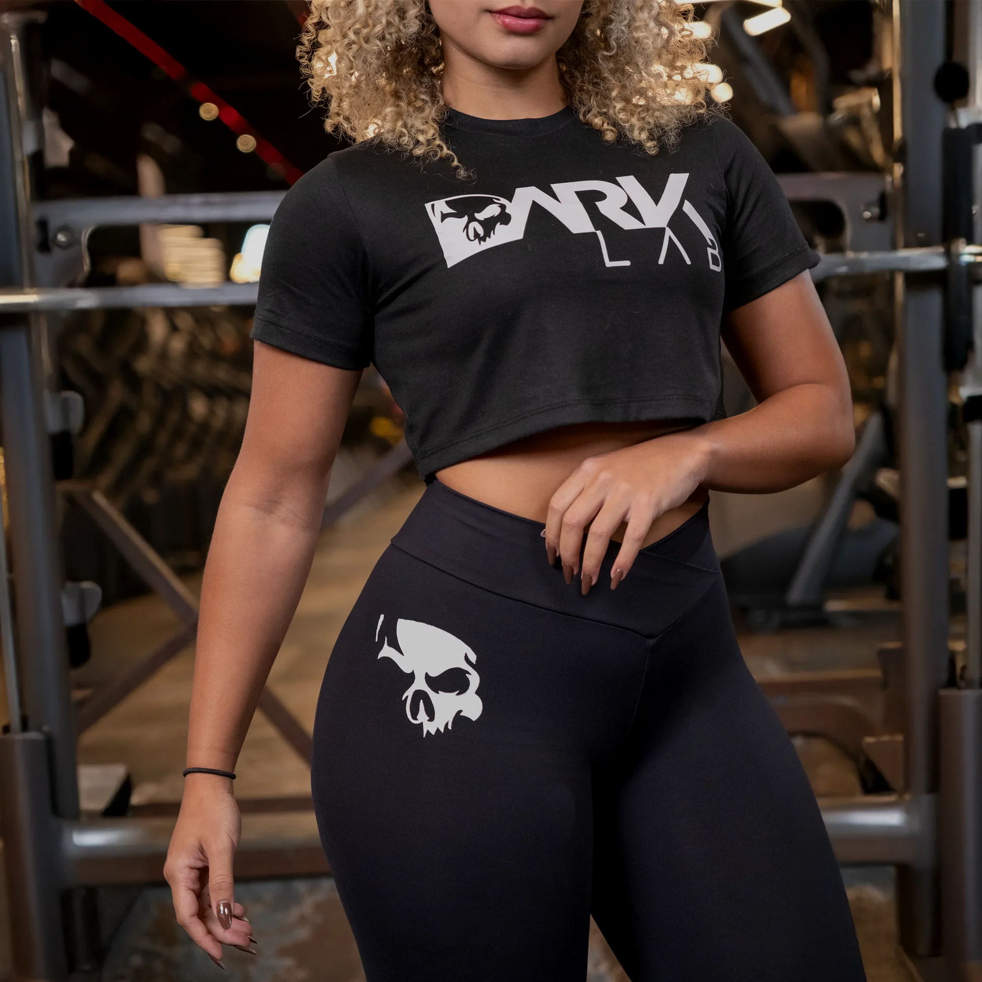Mulher em academia usando top cropped fitness preto e legging preta com logotipo da Dark Labs, em pé diante de equipamentos de musculação.
